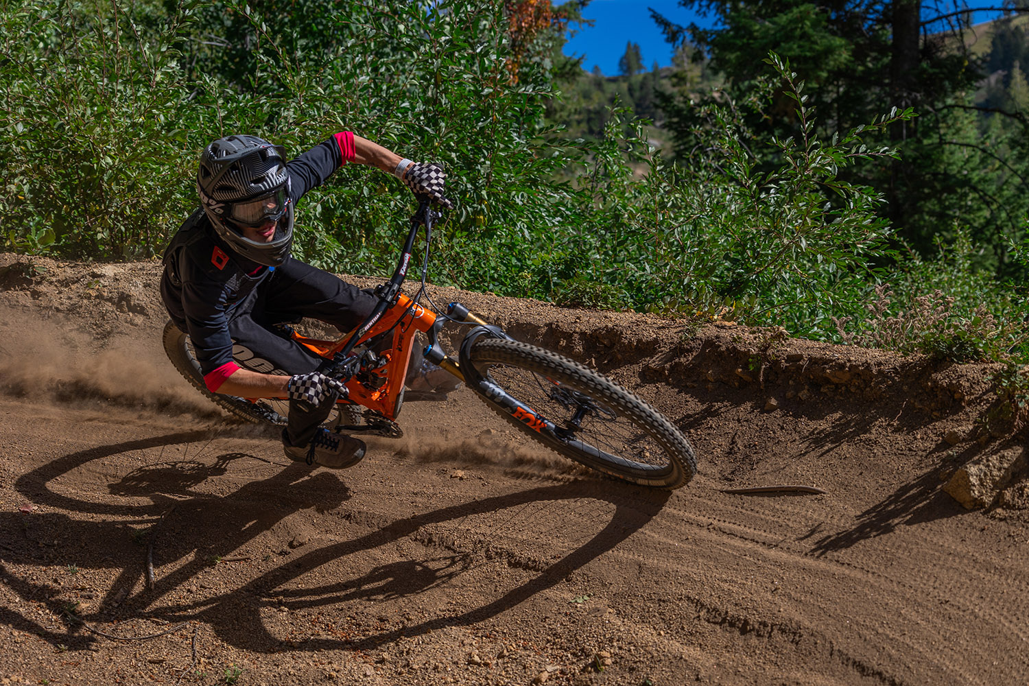 Mountain biker in a full-face helmet leans sharply into a dirt berm, kicking up dust while riding a downhill trail surrounded by green forest.