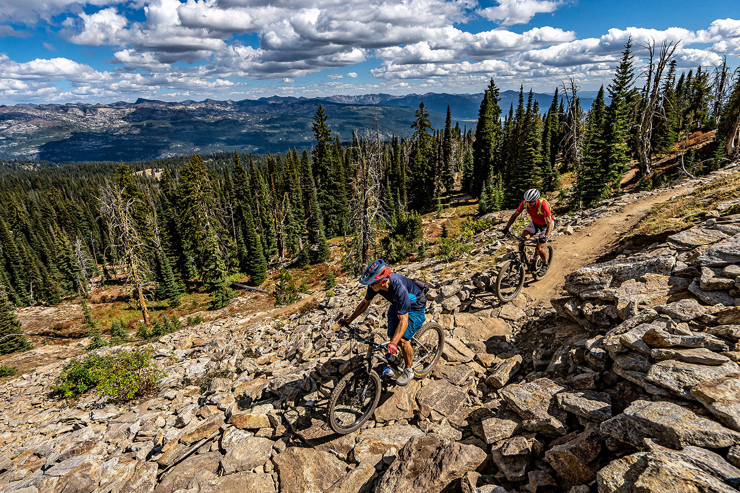 Two mountain bikers ride a rocky alpine singletrack trail above a forested landscape, with mountain views and dramatic clouds in the distance.