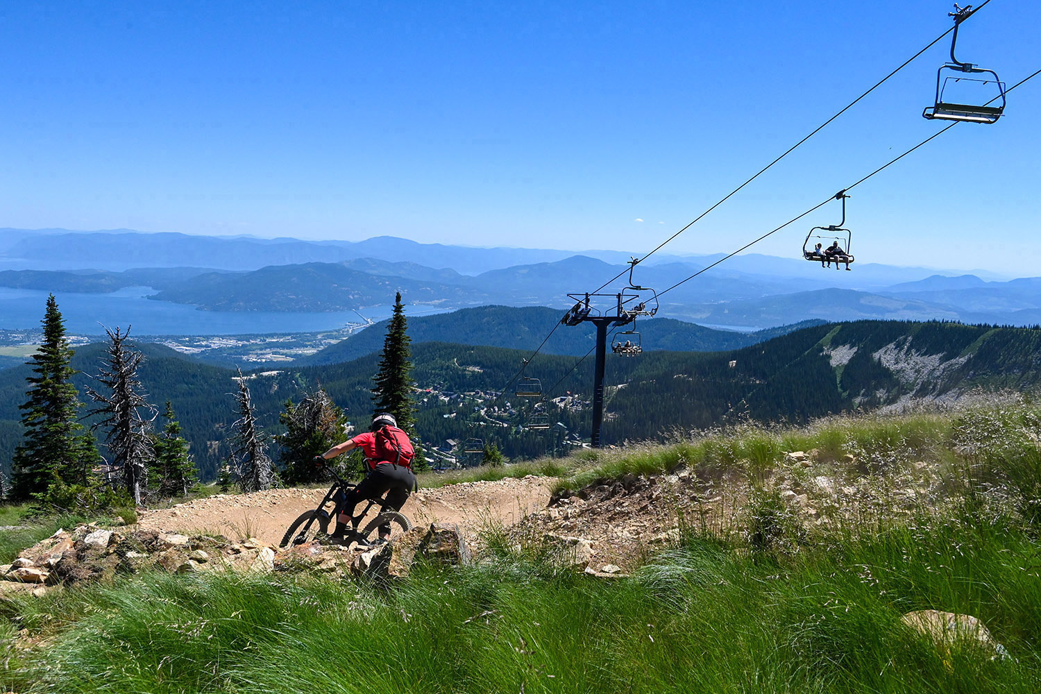 Mountain biker descends a rocky trail at Schweitzer Mountain beneath a chairlift, with Lake Pend Oreille and forested ridgelines visible in the distance.