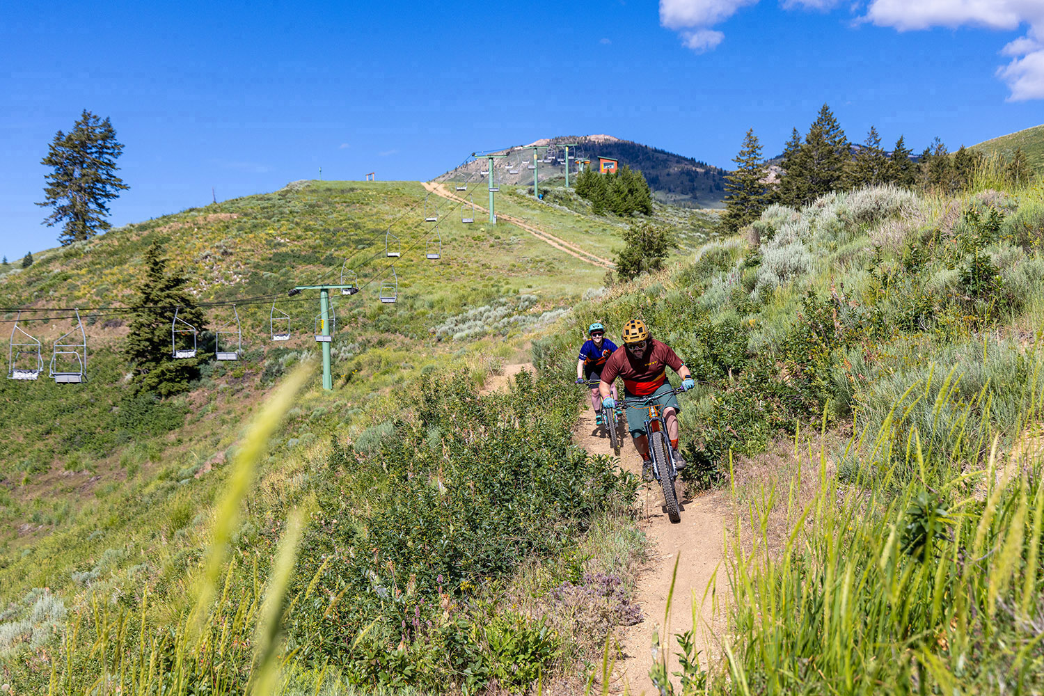 Two mountain bikers ride a narrow singletrack trail through sagebrush-covered hillsides at Soldier Mountain, with chairlift towers and mountain terrain in the background.