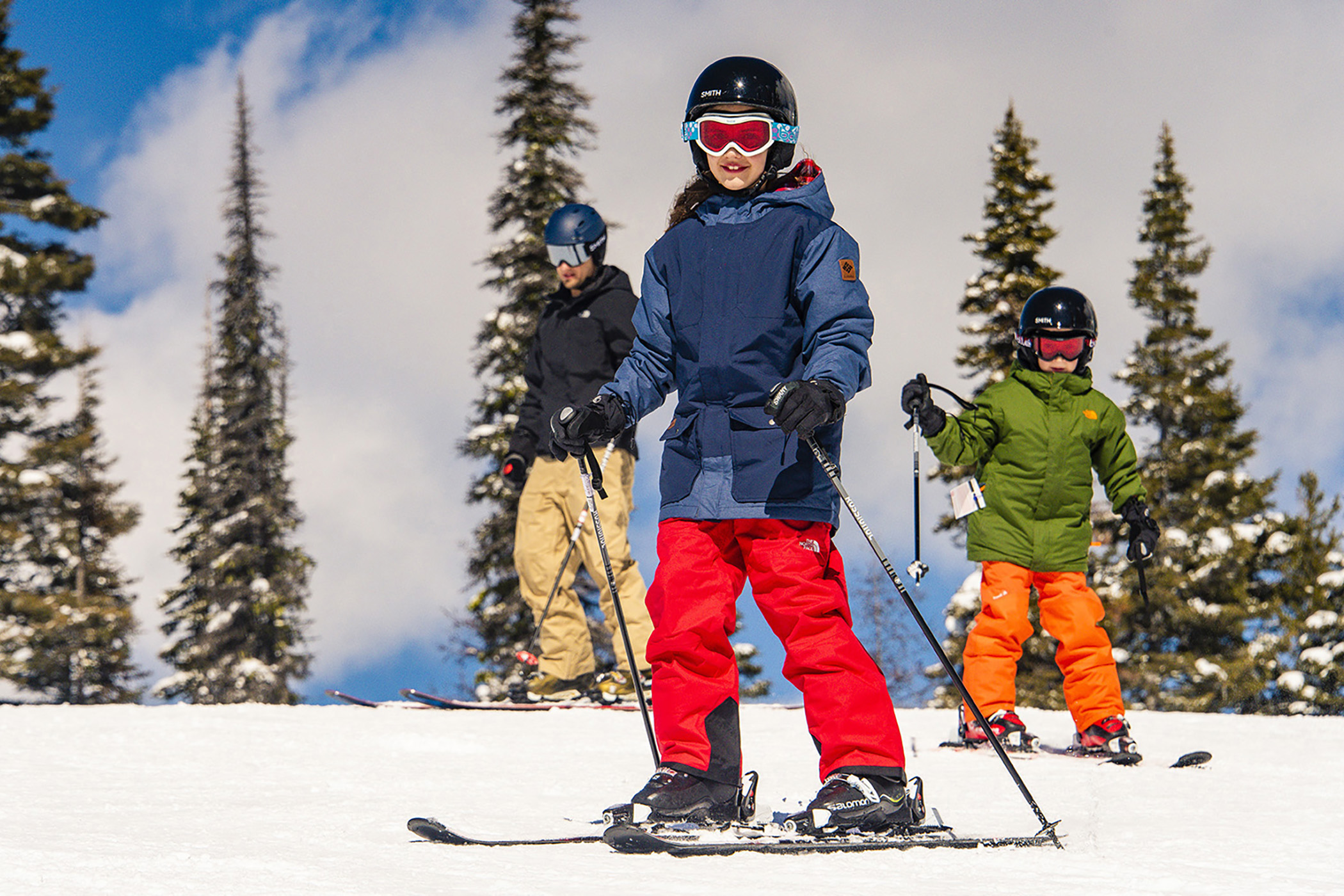 Three skiers carving down a snowy slope surrounded by Idaho evergreens, capturing the fun and confidence‑building winter adventures offered through the Ski Idaho Passport program.