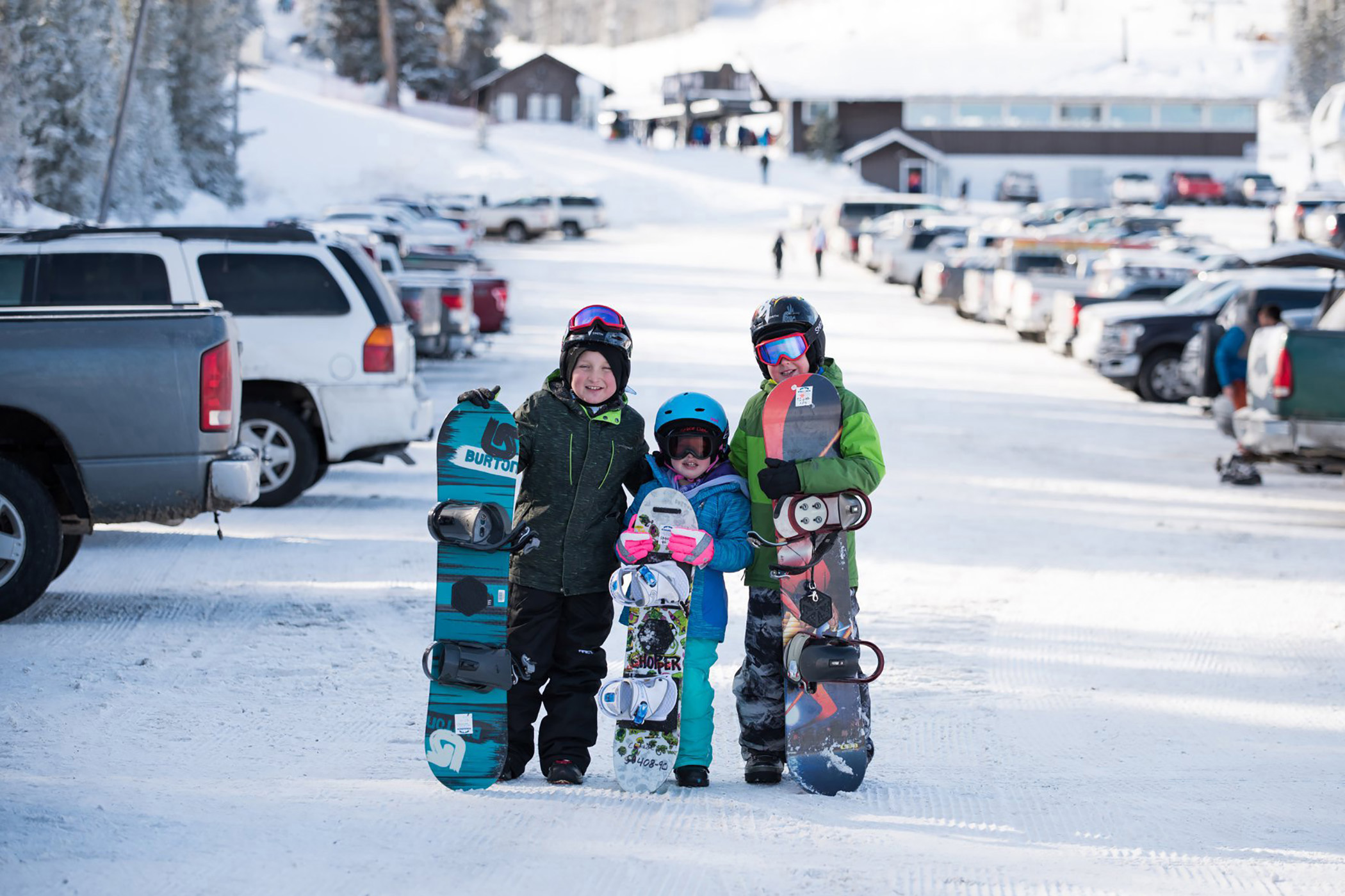 Three children in helmets and winter gear holding snowboards in a ski‑resort parking lot, reflecting the excitement and independence young riders gain through the Ski Idaho Passport program.