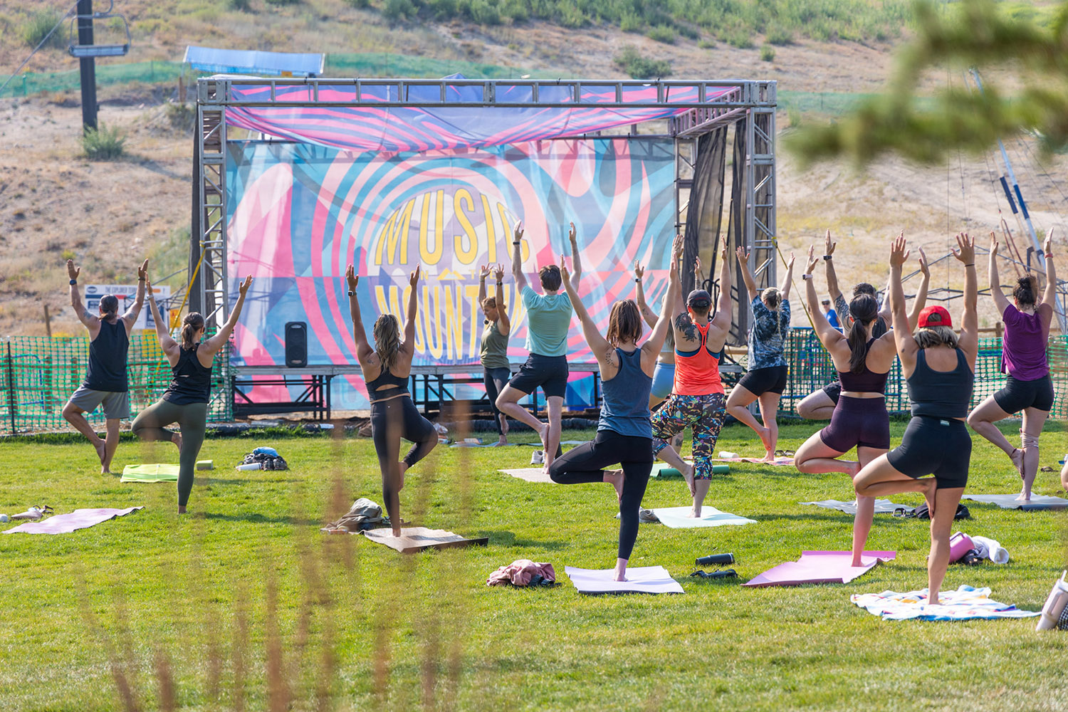 Participants practice Yoga on the Mountain at Bogus Basin in Idaho, balancing in a group pose on a grassy hillside with a Music on the Mountain stage banner in the background.
