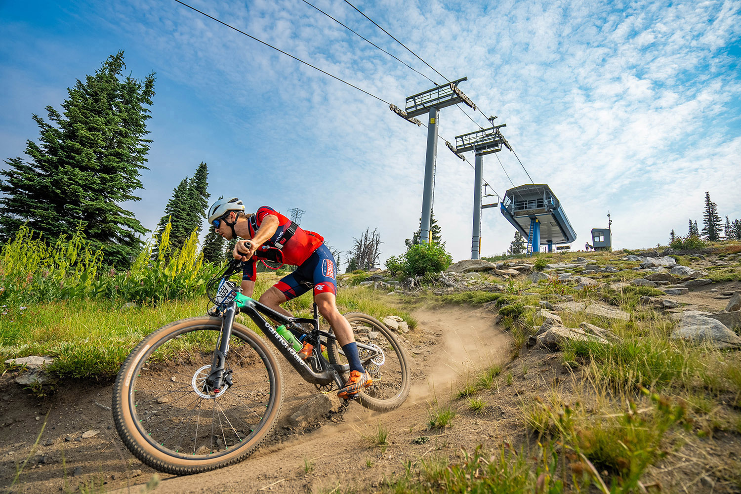 Mountain biker rides a downhill trail at Brundage Mountain Resort in McCall, Idaho, kicking up dust with ski lift towers and alpine terrain in the background.