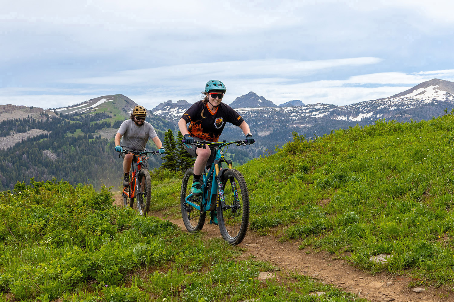 Two mountain bikers climb a singletrack trail at Grand Targhee Resort near Driggs, Idaho, with alpine meadows, evergreen forests, and snow‑dusted peaks in the background.