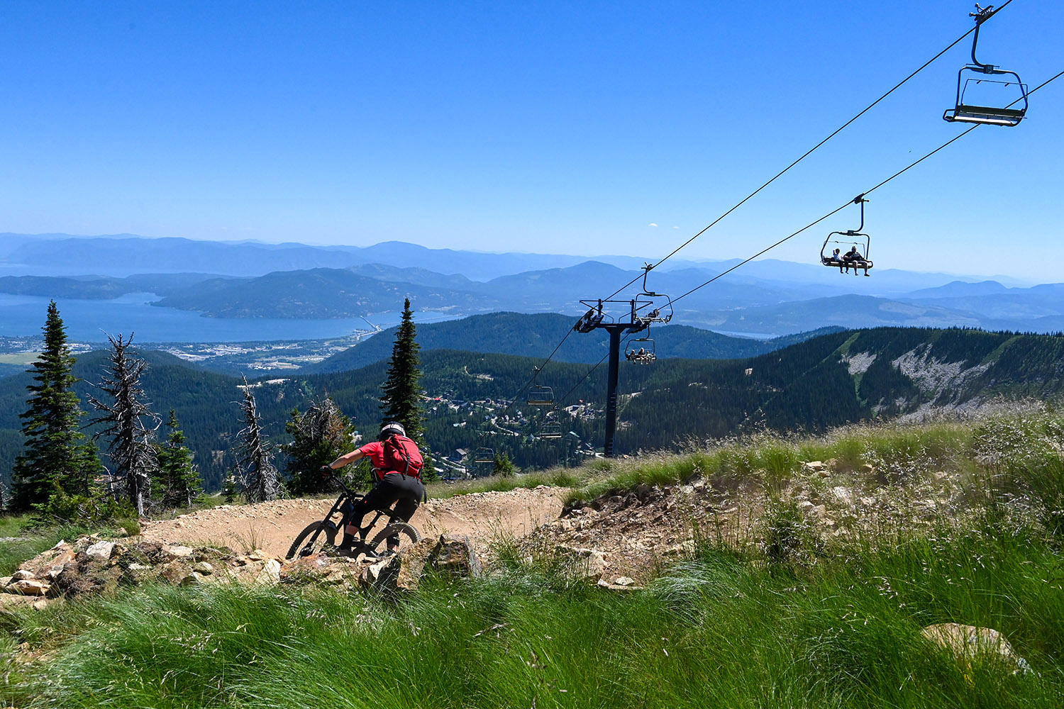Mountain biker rides a downhill trail at Schweitzer in North Idaho, with a scenic chairlift, forested mountains, and Lake Pend Oreille visible in the distance.
